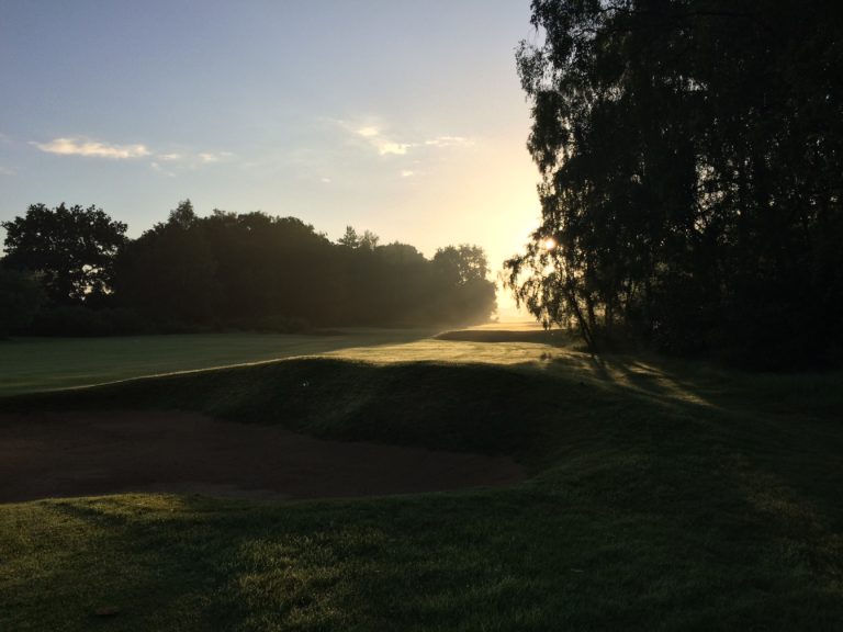 A golf green at sunrise with mature trees along the edge of the fairway
